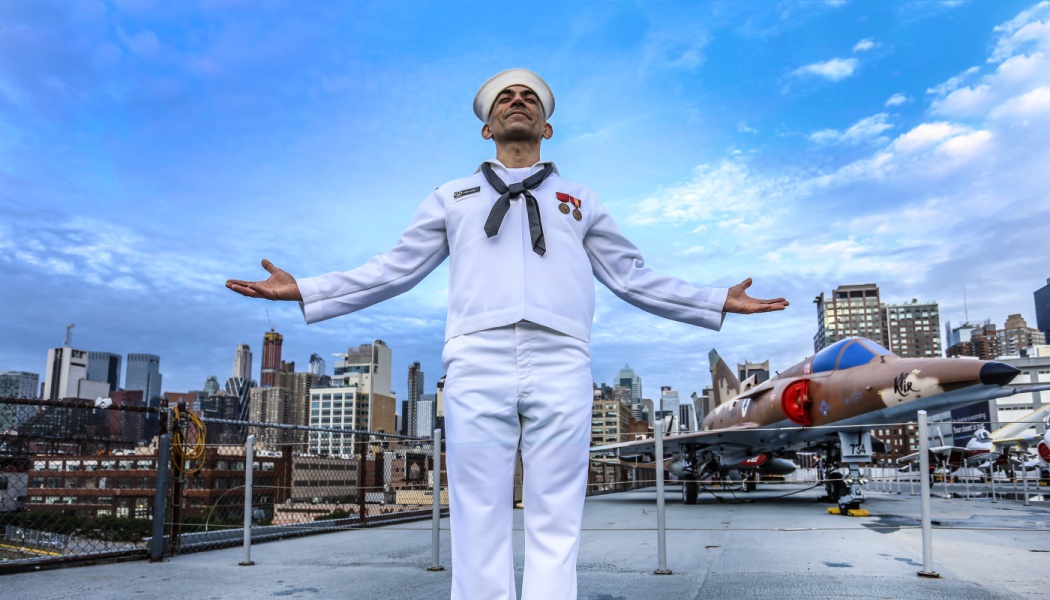 A smiling sailor in a white uniform stands with arms wide open on an aircraft carrier deck near a city skyline.