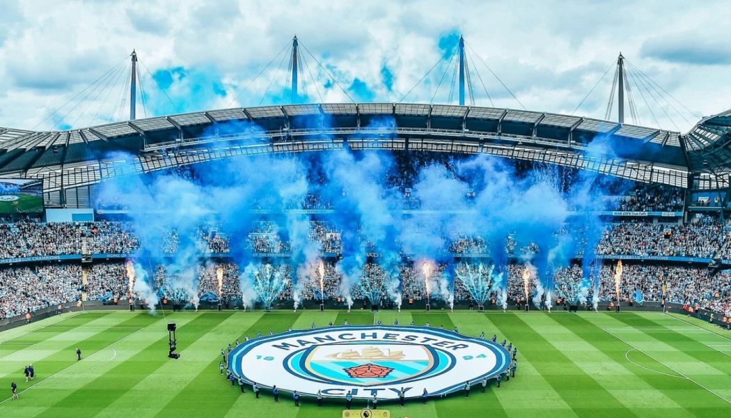 Blue smoke flares and pyrotechnics fill Etihad Stadium behind a giant Manchester City crest displayed on the pitch.