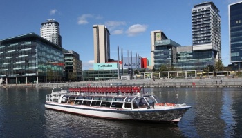 A white passenger boat with red top-deck seats cruises past modern waterfront buildings on a sunny day.