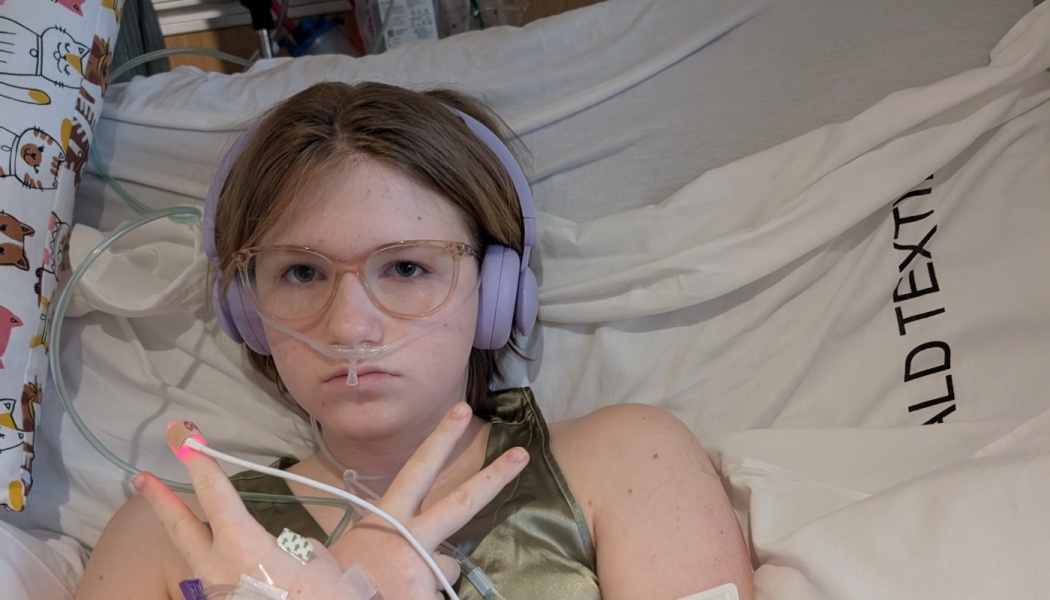 Young person in bed wearing purple headphones, glasses, and a nasal cannula, making a peace sign.