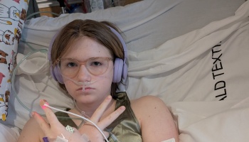 Young person in bed wearing purple headphones, glasses, and a nasal cannula, making a peace sign.