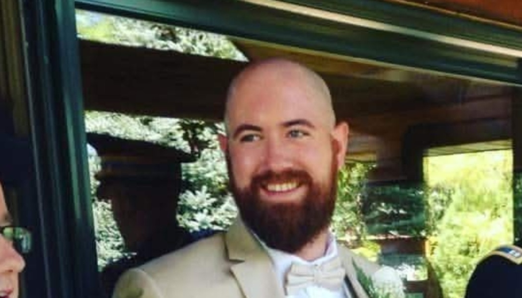 A smiling bald man with a full beard wearing a light tan suit and beige bow tie.