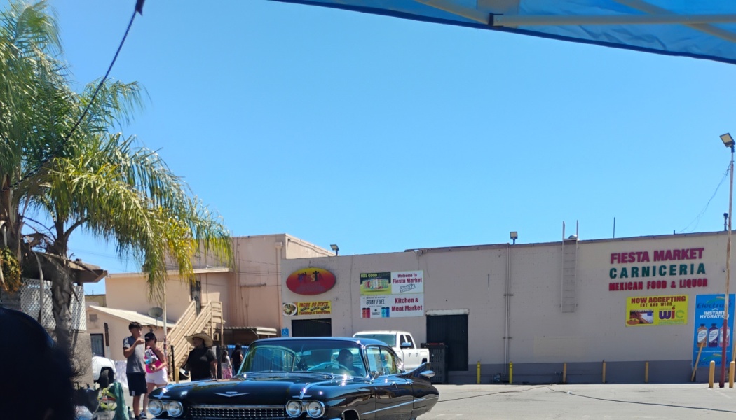 A classic black car parked in front of the Fiesta Market Carniceria on a clear, sunny day.