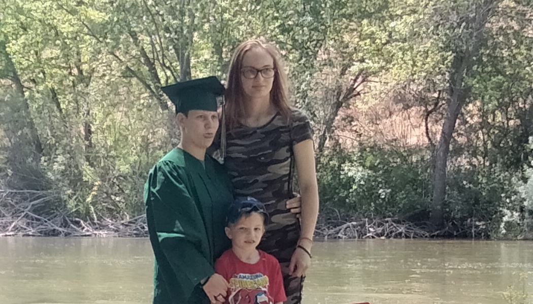 A graduate in a green cap and gown poses outdoors near a river with a woman and a young boy.
