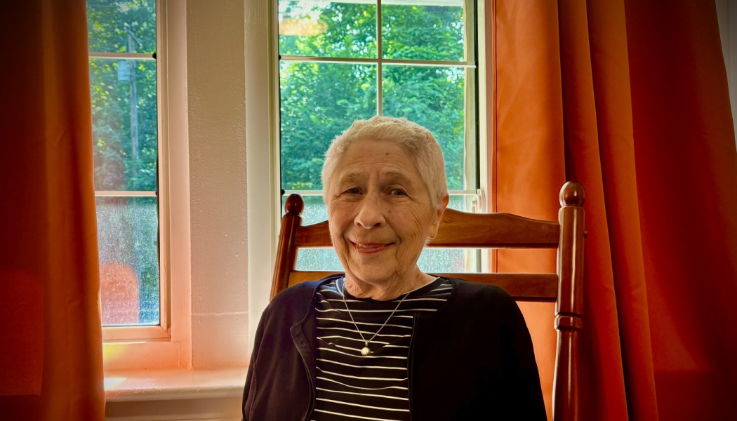 An elderly woman with short grey hair smiles in a wooden chair in front of a window with orange curtains.