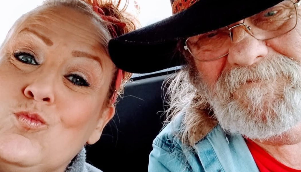 Close selfie of a woman making a kissy face next to an older bearded man wearing glasses and a black hat.