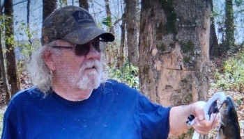 Older bearded man in a camo cap and glasses holding a freshly caught fish in the woods.