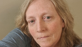 Close-up portrait of a fair-skinned woman with light hair and freckles looking at the camera with a somber expression.