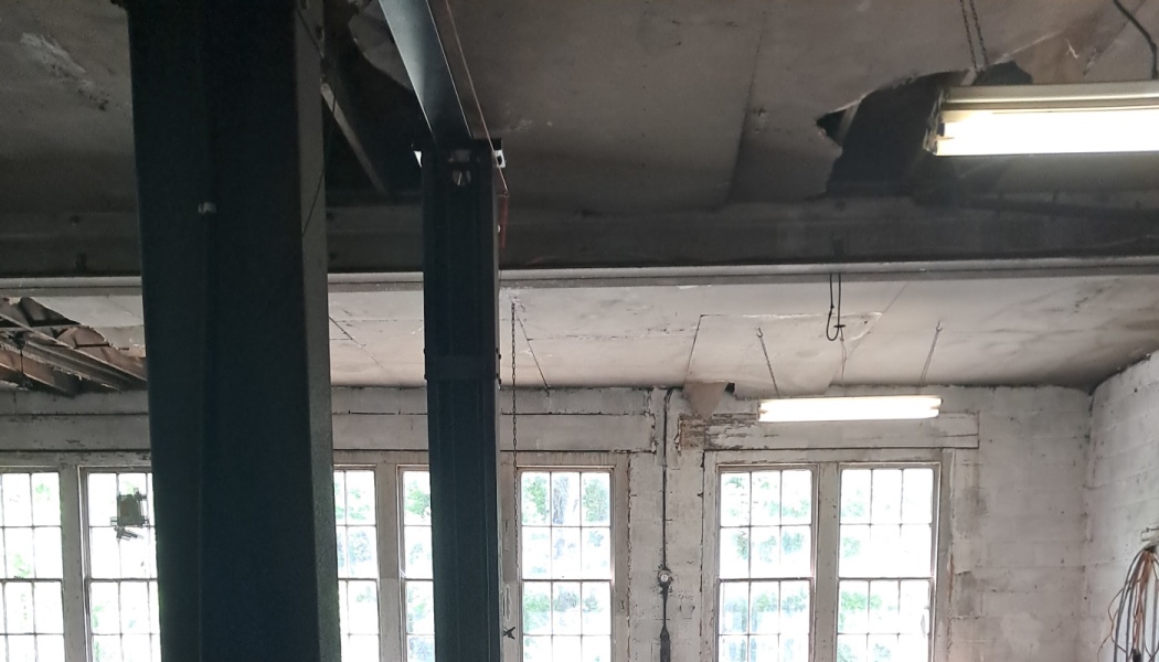 Severe ceiling damage with large holes and hanging lights inside an old workshop with multi-pane windows.