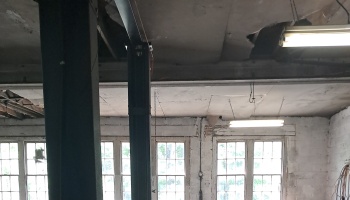 Severe ceiling damage with large holes and hanging lights inside an old workshop with multi-pane windows.