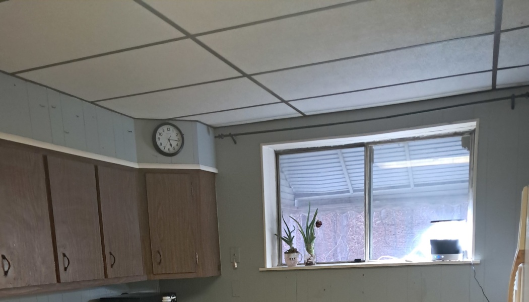Dated kitchen corner with brown cabinets, a drop ceiling, wall clock, and small potted plants on a window sill.