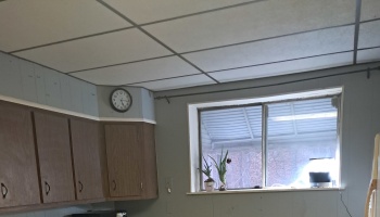 Dated kitchen corner with brown cabinets, a drop ceiling, wall clock, and small potted plants on a window sill.
