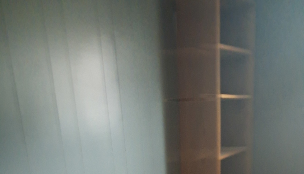 An empty wooden bookshelf stands against a wall in a blurry, dimly lit room.