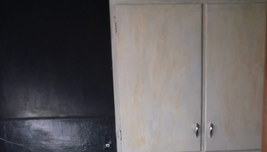 Worn, discolored off-white kitchen cabinets with silver handles mounted next to a dark black wall.