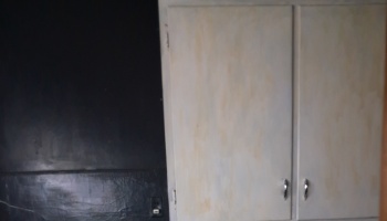 Worn, discolored off-white kitchen cabinets with silver handles mounted next to a dark black wall.
