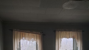 Two windows with off-white lace curtains hanging crookedly in a dim room with ceiling tiles.