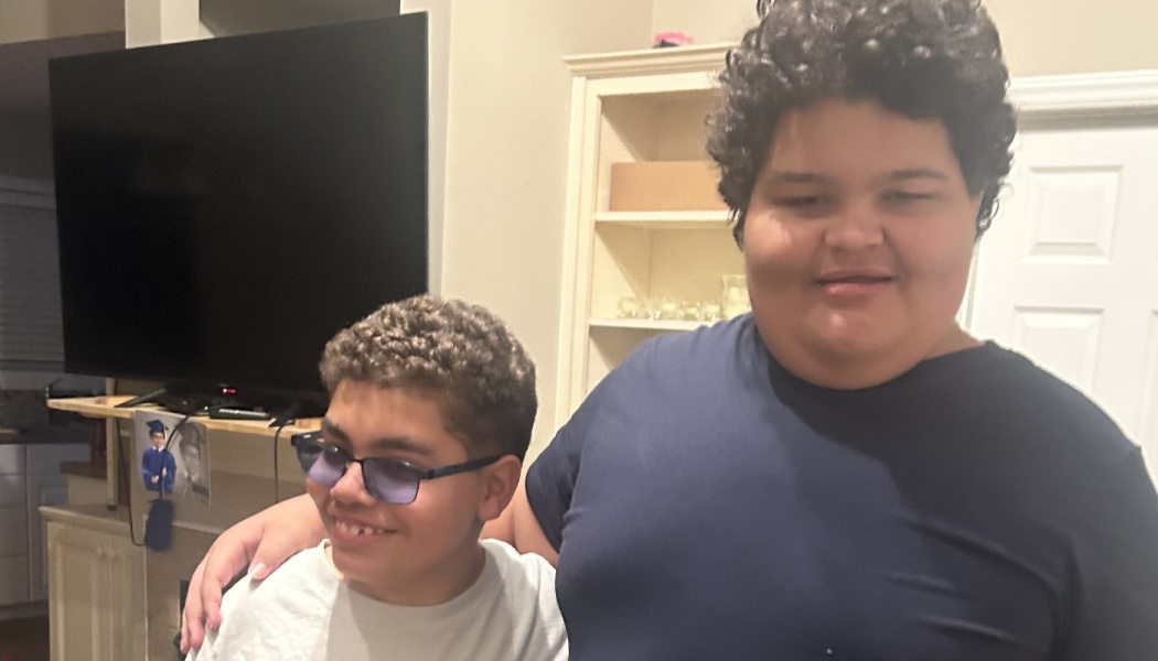 Two smiling boys with curly hair pose together; the older boy puts his arm around the younger boy wearing tinted glasses.