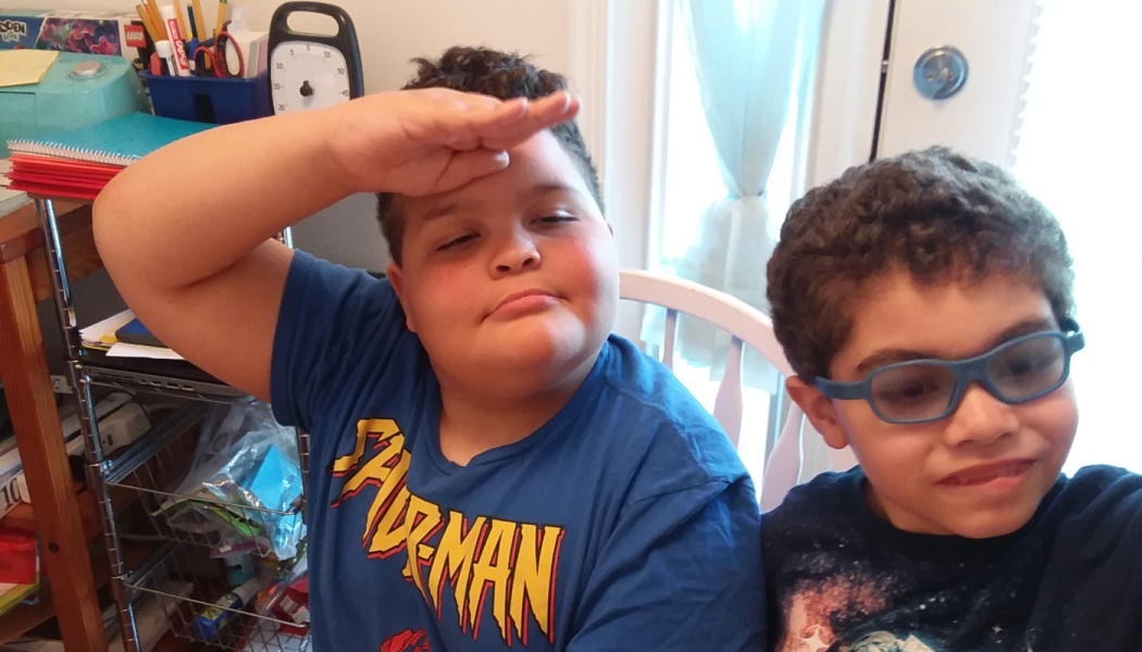 Two young boys sitting together; one in a blue Spider-Man shirt saluting and the other wearing blue glasses.