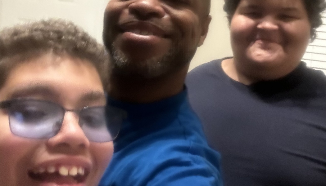 Close-up selfie of a smiling man and two happy children posing together, one wearing glasses.