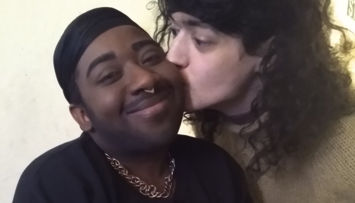 A person with dark curly hair kisses the cheek of a smiling Black person wearing a durag and chain.