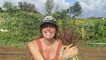 A smiling farmer holds a large bundle of freshly harvested purple carrots in a lush garden field.