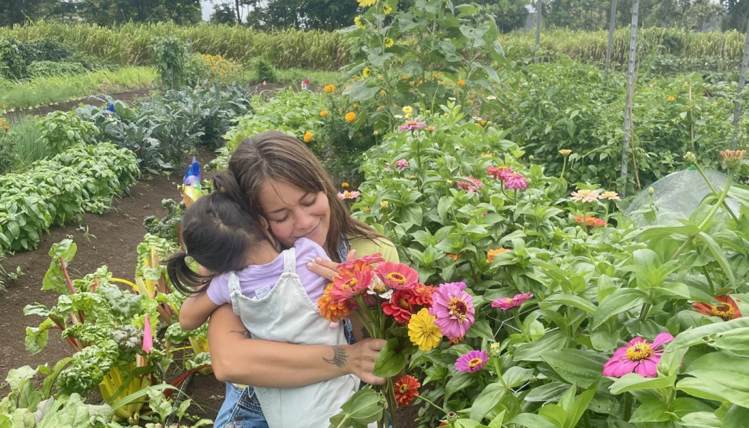 A smiling woman hugs a young child while holding a bouquet of colorful flowers in a lush garden.