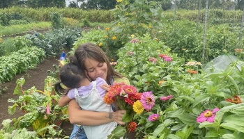 A smiling woman hugs a young child while holding a bouquet of colorful flowers in a lush garden.