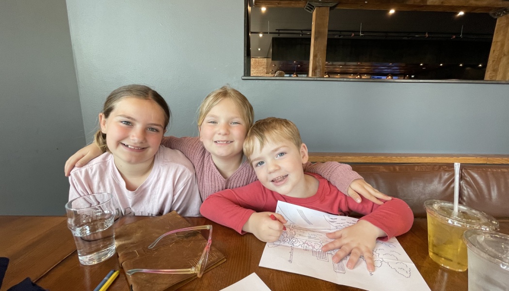 Three smiling young children sitting close together at a restaurant table with a coloring sheet.