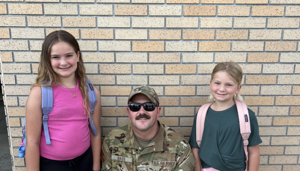A U.S. Air Force soldier poses with two young daughters wearing backpacks against a light brick wall.