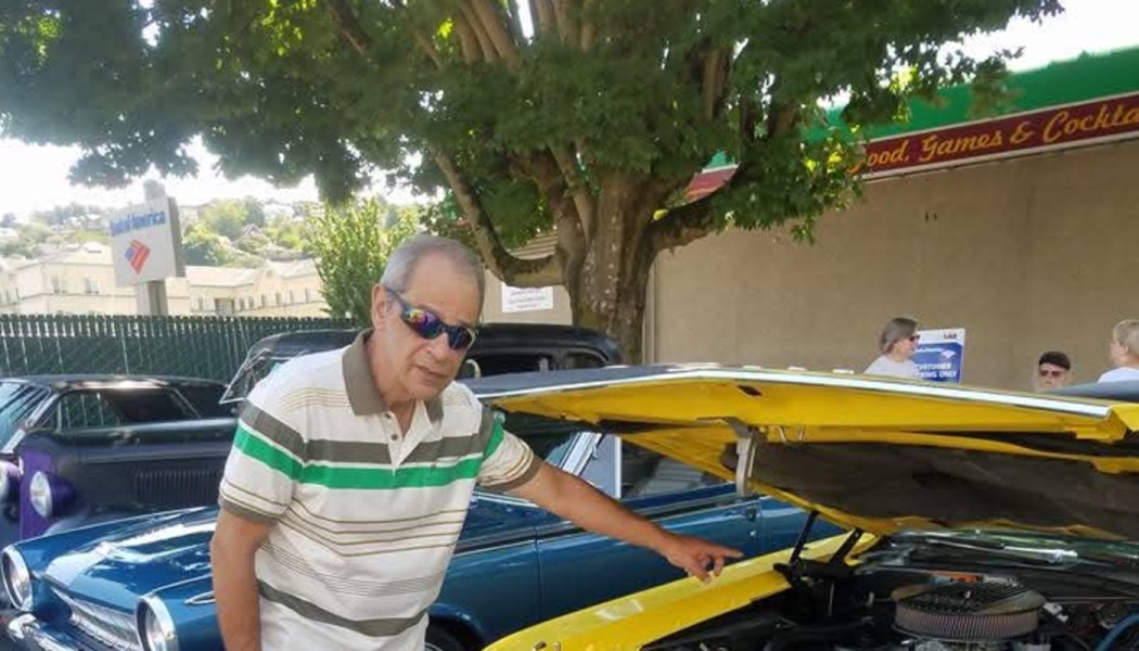 A man in sunglasses and a striped shirt points to the engine under the open hood of a classic yellow car.