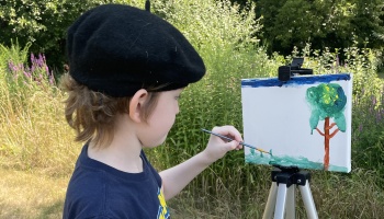 Young child wearing a black beret painting a tree on a canvas easel outdoors.