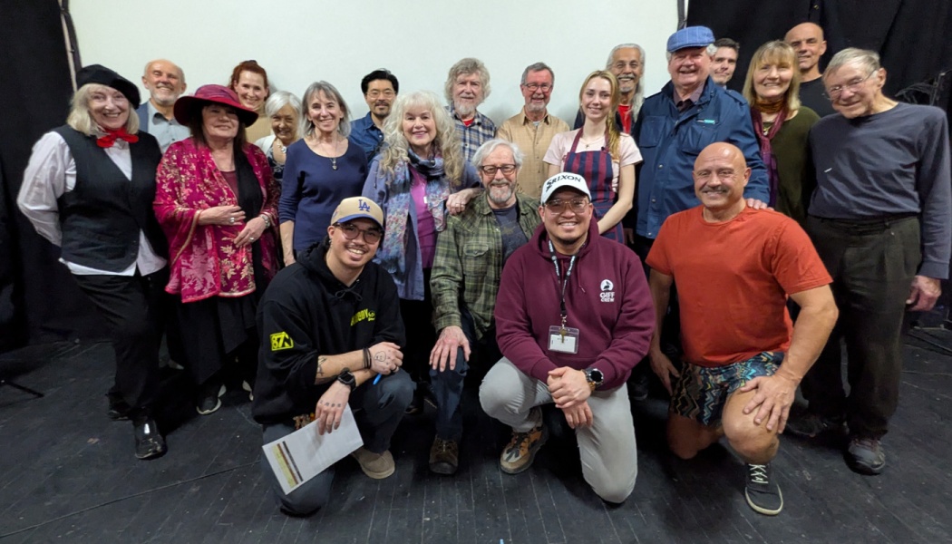 A diverse group of actors and crew members of all ages smiling together on stage.
