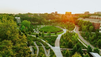 Aerial view of a vibrant green park with winding pathways, dense trees, and a golden sunset shining over buildings.