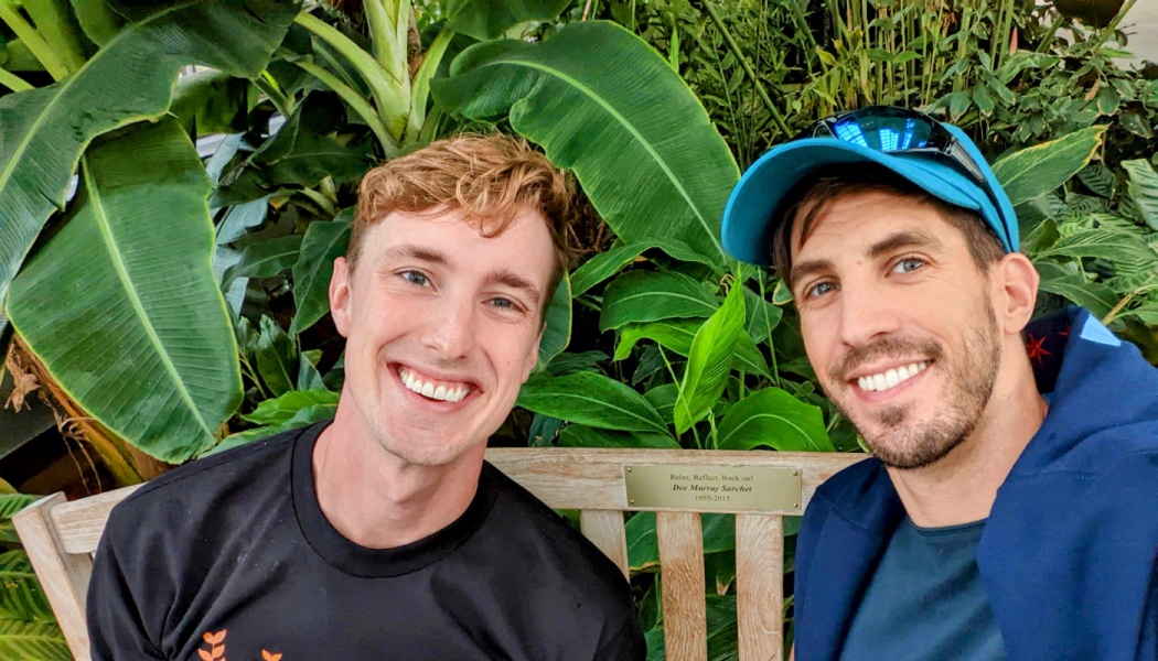 Two smiling young men sit together on a wooden bench surrounded by lush green tropical plants.