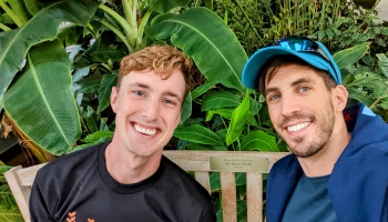 Two smiling young men sit together on a wooden bench surrounded by lush green tropical plants.