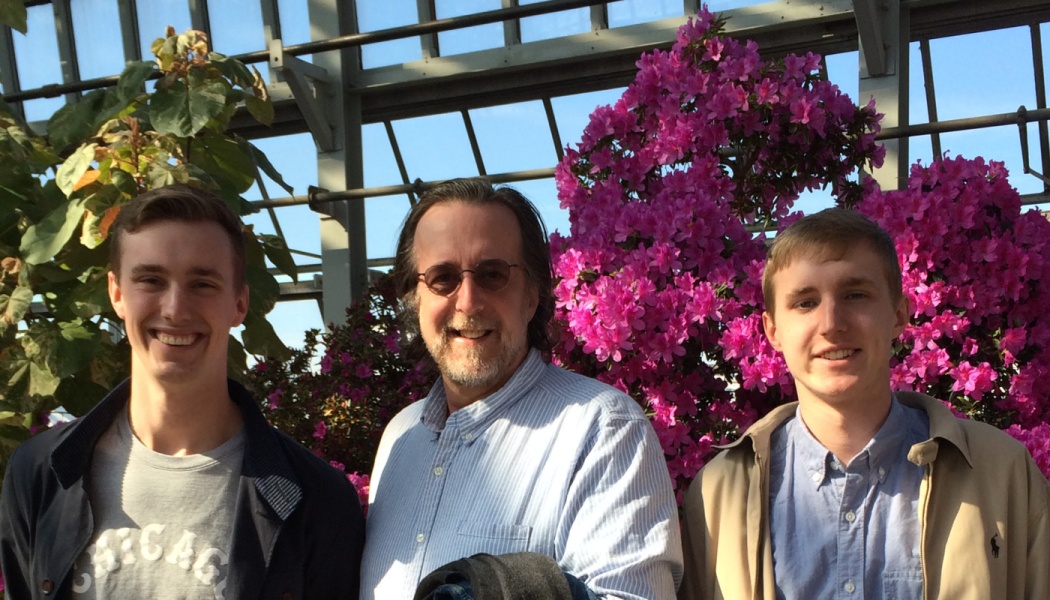 Three smiling men stand in a greenhouse surrounded by vibrant pink flowers and green foliage.