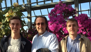 Three smiling men stand in a greenhouse surrounded by vibrant pink flowers and green foliage.