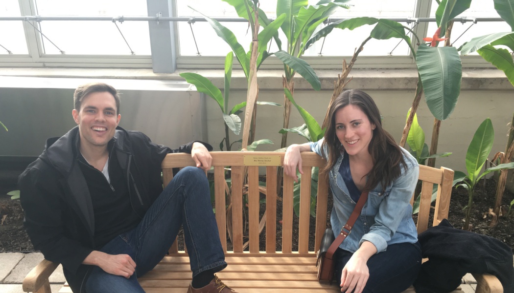 A smiling man and woman sit on a wooden bench surrounded by large tropical plants.
