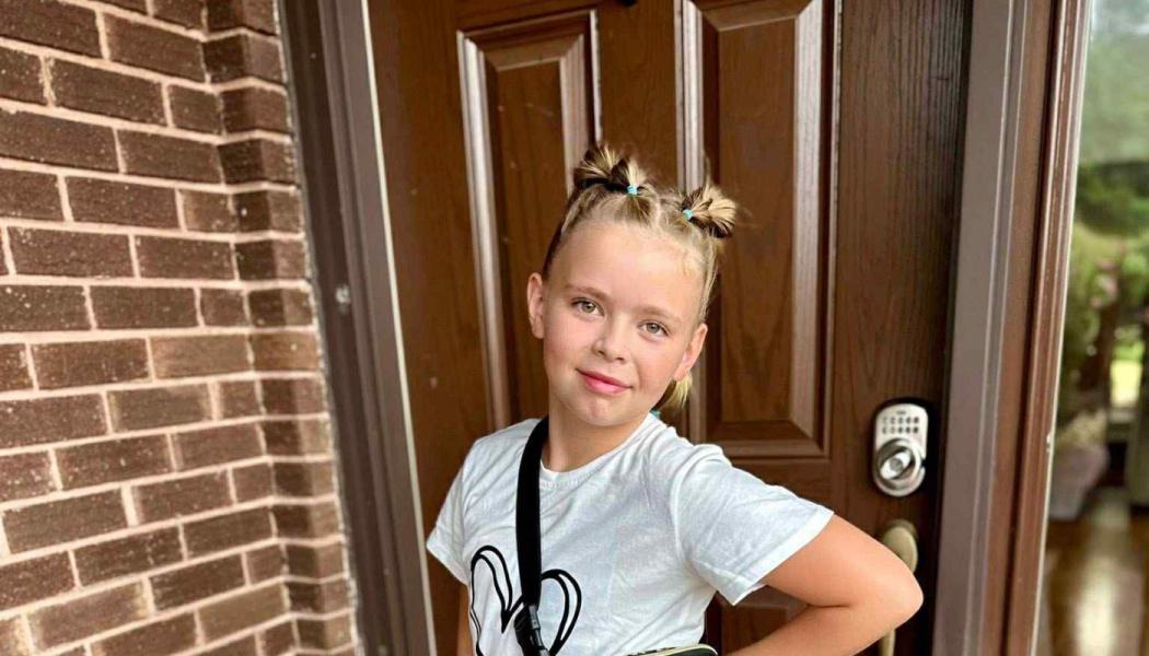 Young girl with blonde hair in space buns smiling in front of a wooden door.