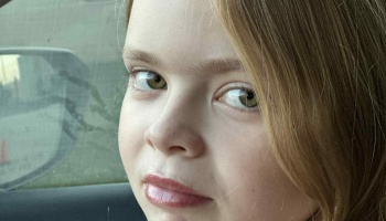 Close-up portrait of a young girl with light hair and hazel eyes looking at the camera inside a car.