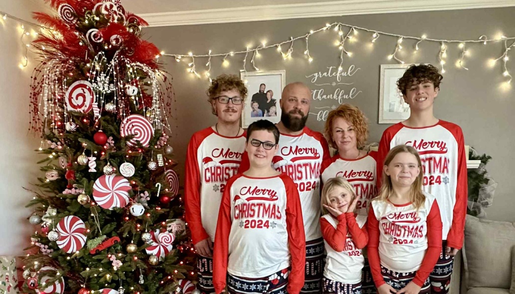 A family of seven wearing matching red and white holiday pajamas poses beside a decorated Christmas tree.
