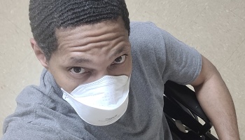 A man wearing a white N95 mask and grey shirt looking up while seated in a wheelchair.