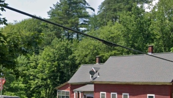 Red building with a grey roof surrounded by dense green forest, with utility lines extending across the foreground.