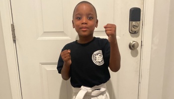 Young boy in a martial arts uniform with a white belt posing proudly with raised fists.
