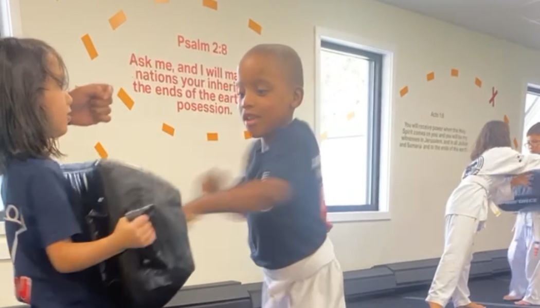 A young boy in a martial arts uniform punches a pad held by a classmate during practice.