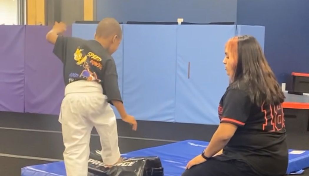 A young boy in white martial arts pants practices on a pad while an instructor watches.