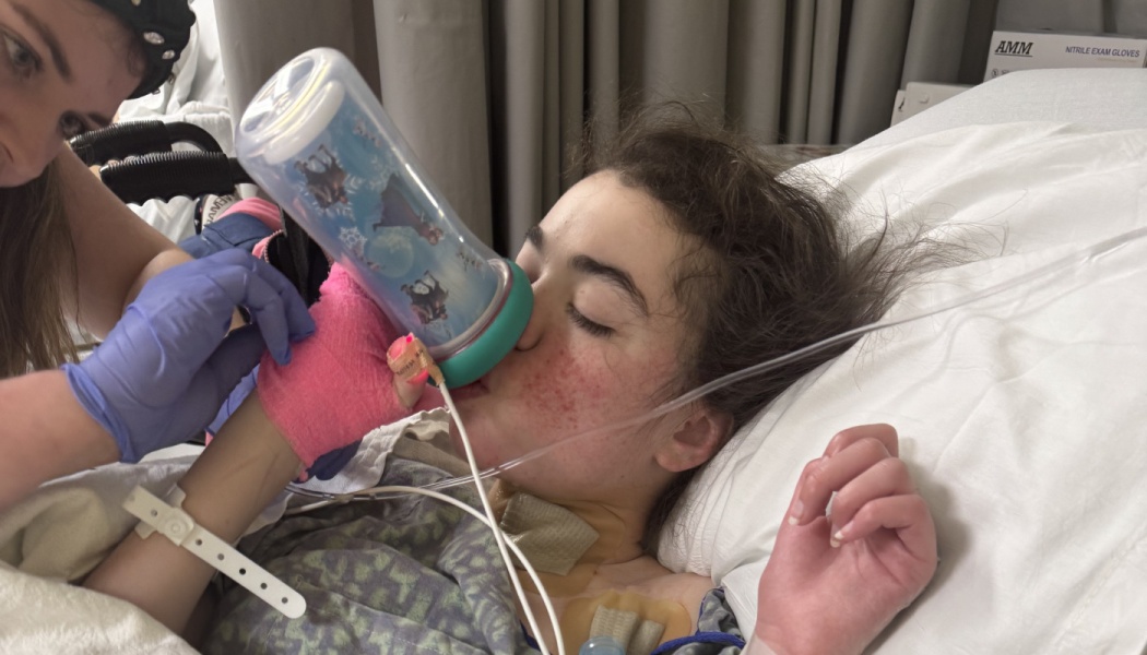 A caregiver in purple gloves helps a young patient lying in a hospital bed drink from a bottle.