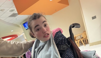 A young girl in a wheelchair looking toward the camera, wearing a heart-patterned jacket and grey towel.
