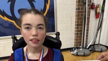 Young girl sitting in a wheelchair wearing a red, white, and blue medal inside a gymnasium.