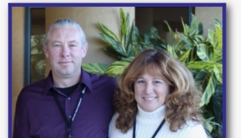 A grey-haired man in a purple shirt and a smiling woman in a white turtleneck posing together near green plants.